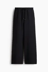 Pantalón negro de corte holgado y tiro alto, confeccionado en tejido plumeti (textura con pequeños puntos en relieve). Presenta cintura elástica fruncida con volados y cordón ajustable, además de bolsillos discretos en las costuras laterales.