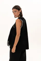 Chaleco negro de gamuza con diseño sin mangas, decorado con ojales metálicos plateados a lo largo de los bordes frontales y sisas.