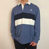 Buzo tipo polo de rugby con diseño de bloques de color en azul, azul marino y blanco. Presenta cuello camisero blanco con botones, mangas largas con puños elásticos y un pequeño logo bordado en el pecho.