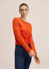 Camiseta de manga larga color naranja, con cuello redondo y diseño entallado. Presenta un detalle fruncido central y costuras decorativas en el frente.