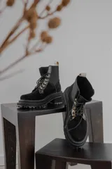 Borcego de gamuza color chocolate con paneles laterales elásticos negros y suela track dentada. Presenta sistema de cordones con ganchos metálicos y tirador en la parte trasera.