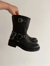 Bota de caña media en eco cuero negro, con diseño estilo biker que incluye correas con hebillas metálicas y herrajes plateados en el tobillo. Cuenta con suela plana y punta redondeada.