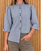 Camisa de denim y algodón a rayas azules y blancas, con cuello alto con volados, mangas tres cuartos abullonadas con puños con volados y cierre frontal de botones oscuros.