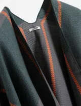 Ruana de punto color verde oscuro con franjas verticales en diseño de zigzag color naranja. Presenta un diseño abierto y holgado sin mangas, confeccionada íntegramente en poliéster.