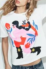 Remera blanca de corte oversize con cuello redondo y mangas cortas. Presenta un estampado artístico a color en el frente que ilustra una figura femenina estilizada con botas estilo western.