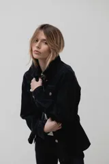 Chaqueta de denim negro con corte entallado en la cintura, cierre frontal con botones metálicos y bolsillos en el pecho.