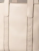 Cartera tote Calvin Klein de color beige, confeccionada en suave piel sintética, con diseño estructurado, asas fijas y compartimentos exteriores en los laterales. Presenta detalles de cinta con logo en jacquard y logo metálico grabado en la parte delantera.