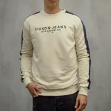 Buzo color beige con mangas largas y cuello redondo. Presenta una banda azul marino a lo largo de las mangas y un estampado frontal con la leyenda "Duxon Jeans Los Angeles Est 1982".
