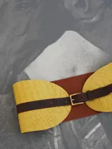 Cinturón de diseño escultórico con base acolchada en color amarillo y correa de cuero marrón con hebilla metálica dorada.