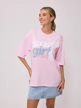 Remera de corte oversize color rosado, con cuello redondo y mangas cortas. Presenta un estampado frontal con la frase 'surf girl' y el logo de la marca Rusty, acompañado de un diseño floral.
