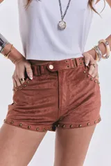Short corto de tiro bajo confeccionado en gamuza elastizada color marrón camel. Presenta detalles de tachas metálicas doradas en los bolsillos delanteros y en los bordes inferiores, además de una faja en la cintura con hebillas metálicas laterales.