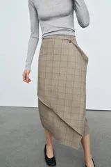 Falda midi de tiro medio con estampado de cuadros en tonos beige y gris. Presenta un diseño cruzado con un volante decorativo en el delantero y cierre con botón.