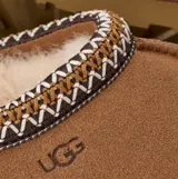 Zuecos UGG modelo Tasman II, color tostado, confeccionados en gamuza con forro interior de piel de oveja. Presentan un ribete trenzado característico en el cuello y suela de goma resistente.