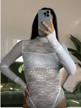 Body de encaje blanco con cuello alto y mangas largas de microtul. Presenta un diseño entallado con detalles de puntilla en los puños y en el borde inferior, combinando transparencias y texturas delicadas.