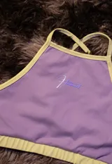 Top deportivo color lila con ribetes en amarillo claro. Presenta tirantes finos y un logo estampado en el centro en tono lila más oscuro.
