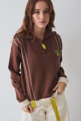 Buzo de tejido de punto color marrón con cuello alto y cierre frontal parcial. Presenta rayas verticales finas en color claro, puños y ruedo en tejido acanalado color crema, y detalles en color verde lima en el tirador del cierre, el cordón de ajuste del ruedo y el parche bordado en el pecho.