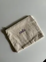 Neceser de tela color crudo con cierre superior y texto 'cada.' bordado en lila.