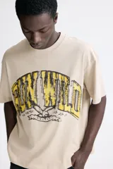Remera de algodón en color beige con cuello redondo acanalado, hombros caídos y corte holgado. Presenta un estampado frontal con la frase 'RUN WILD' en tonos amarillos y negros con efecto desgastado.