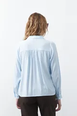 Camisa de manga larga en color celeste, con diseño de corte relajado, cuello clásico y detalle de tablas verticales en el frente.