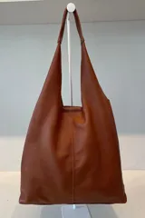 Bolso tipo tote de gran capacidad, confeccionado artesanalmente en cuero vacuno color marrón. Presenta un diseño minimalista con costura central vertical, fuelle lateral ajustable con cierre y placa metálica con logo en la parte inferior.