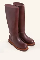 Bota de caña alta confeccionada en cuero vacuno suave, con diseño minimalista y suela de goma robusta. Presenta costuras visibles en la zona del empeine y un taco bajo cuadrado.