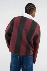 Polo de manga larga con rayas verticales en borgoña y negro, cuello de contraste blanco y cierre frontal parcial con botones ocultos. Presenta diversos bordados y parches decorativos en el pecho.