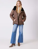 Campera puffer color marrón con capitoneado horizontal. Tiene cuello alto y capucha ajustable con cordones, con el interior del cuello en textura aterciopelada negra. Cierre frontal completo y bolsillos laterales. El corte es ligeramente más largo en la parte posterior con aberturas laterales redondeadas.