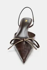Zapato destalonado de mujer, color marrón oscuro con acabado brillante, punta fina y tacón alto y fino. Presenta un detalle de lazo con herraje dorado en la parte delantera y tira elástica en el talón.