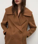 Campera pesada de estructura firme, color marrón, con cuello amplio de solapa alargada, cierre cruzado con botones, mangas largas y bolsillos delanteros. Presenta un canesú sobrepuesto en la espalda con pliegue central.