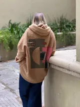 Campera tipo hoodie confeccionada en tejido de corderito color camel, con diseño de collage en la espalda que combina parches de diferentes texturas, colores y estampados geométricos. Presenta capucha y corte holgado.