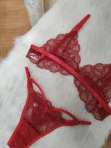 Conjunto de lencería compuesto por un bralette y una tanga, ambos confeccionados en encaje rojo con detalles de tiras elásticas. El bralette presenta un diseño triangular sin aros, mientras que la tanga cuenta con laterales ajustables y diseño de tiras.