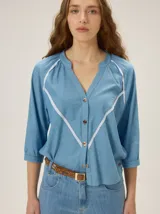 Camisa azul de efecto denim, con escote en V, cuello mao y mangas 3/4. Presenta detalles de ribetes blancos en forma de V en el frente y cierre central de botones.