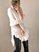 Camisa blanca de tejido suave tipo seda, con mangas tres cuartos y volados irregulares en mangas y ruedo. Tiene un largo ligeramente mayor en la espalda.