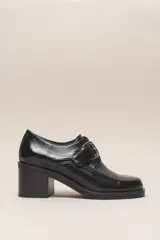 Zapato de cuero vacuno charol negro, con punta redondeada y detalle de perforado en la capellada. Tiene una tira con hebilla metálica y un taco cuadrado de 4 cm.