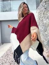 Poncho tejido de punto con diseño de bloques de color en tonos bordó, beige, blanco y negro, con terminación de flecos en el ruedo.