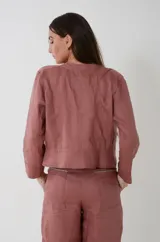 Chaqueta de lino estilo napoleónica color rosa palo, de cuello mao, manga larga y corte a la cintura. Cuenta con cierre frontal parcial de metal y detalle en la pechera con aplicación de cintas y botones decorativos.