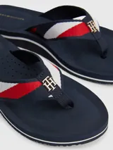 Ojotas de mujer color azul marino con tira de tela trenzada que presenta franjas rojas y blancas, y un dije metálico con el monograma TH en dorado.