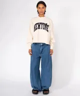 Buzo de algodón color verde oliva con lavado especial, cuello redondo y mangas largas con puños ajustados. Presenta un estampado frontal con la palabra "NEWTONE" en letras negras con efecto puff, y apliques decorativos redondos en el pecho. El ruedo inferior es cortado y deshilachado.