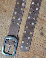 Cinto de cuero marrón con hebilla metálica de estilo envejecido y detalle de tachas metálicas en diferentes tonos en el extremo.