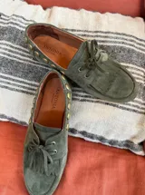 Náuticos de gamuza color verde oliva, con diseño de flecos en la parte frontal, cordones y detalle de tachas metálicas doradas en los laterales. Interior forrado en cuero.