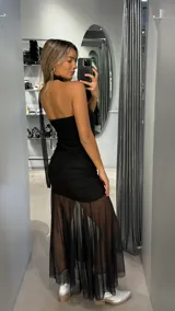 Vestido largo negro de diseño asimétrico con un solo hombro y detalle de choker integrado. Presenta un cuerpo ajustado con frunces laterales y una falda inferior con transparencia en tul, creando un efecto de capas.