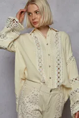 Blusa color crema con mangas largas estilo kimono y detalles de encaje calado (broderie) en el frente y puños. Presenta cuello camisero y cierre frontal con botones plateados.