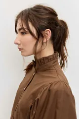 Camisa de manga larga color marrón, con cuello alto con volados, botonadura frontal y detalles calados en las mangas abullonadas.