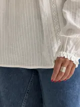 Blusa blanca de corte holgado con cuello redondo, mangas largas con puños elásticos y detalles de puntillas verticales en el frente.
