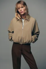 Chaqueta bomber negra con cuello solapa, manga larga abullonada y puños elásticos. Presenta falsos bolsillos de doble vivo en la parte delantera y cierre frontal con cremallera.