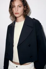 Trench corta de color negro con cuello de solapa, cierre frontal cruzado con botones y mangas largas con trabillas ajustables. Cuenta con bolsillos delanteros de vivo.