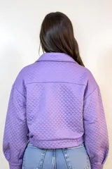 Campera corta de textura capitoneada en color lila, con cuello camisero, cierre frontal con botones y bolsillos con solapa en el pecho. Presenta mangas largas con puños elásticos y diseño holgado.