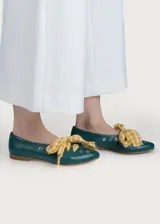 Bailarina de cuero color verde oscuro con detalles de ojales metálicos plateados y lazo desmontable de tela a cuadros amarillos y blancos. Presenta una silueta clásica con punta redondeada y suela plana.
