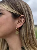 Aro tipo argolla dorada con un dije colgante de disco plano y texturizado. En el cartílago superior se observa un aro tipo cuff liso dorado.