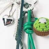 Llavero decorativo con mosquetón en forma de corazón, compuesto por una cadena de perlas, cintas de tela verde, cordones técnicos y un dije tejido a crochet con forma de kiwi.